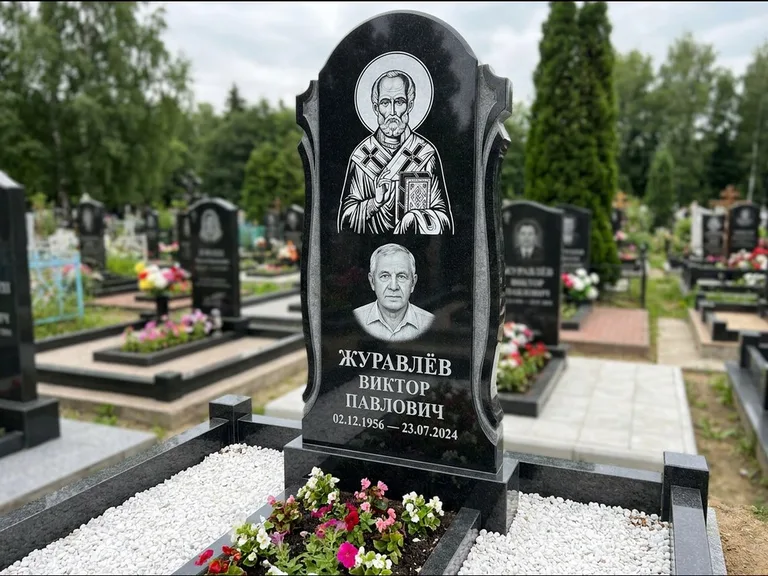 Памятник с гравировкой иконы: стела с образом в полный рост на обороте — гравировка образа Матроны Московской, низкие кусты спиреи в цвету, работа «Лебедева» в Туле